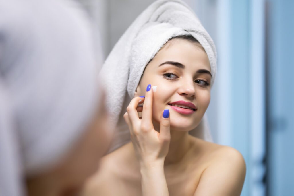 smiling young woman applying cream to face and looking to mirror at home bathroom